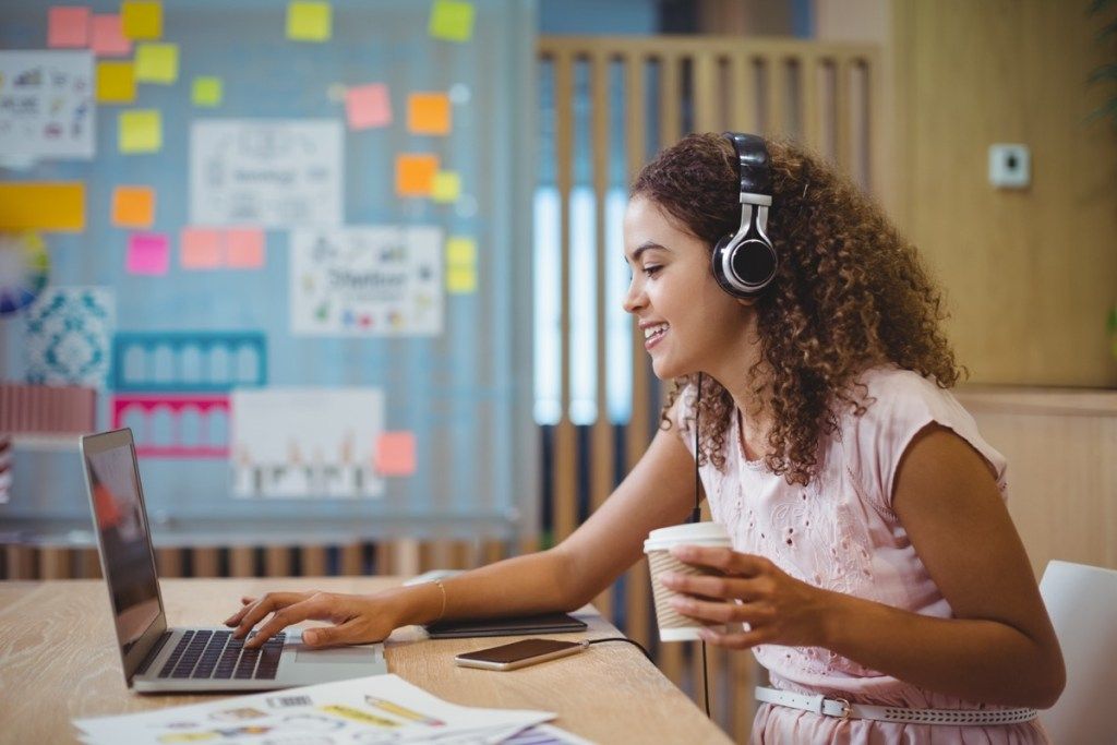 Nainen graafinen suunnittelija kuuntelee laulua käyttäessään kannettavaa tietokonetta toimistossa