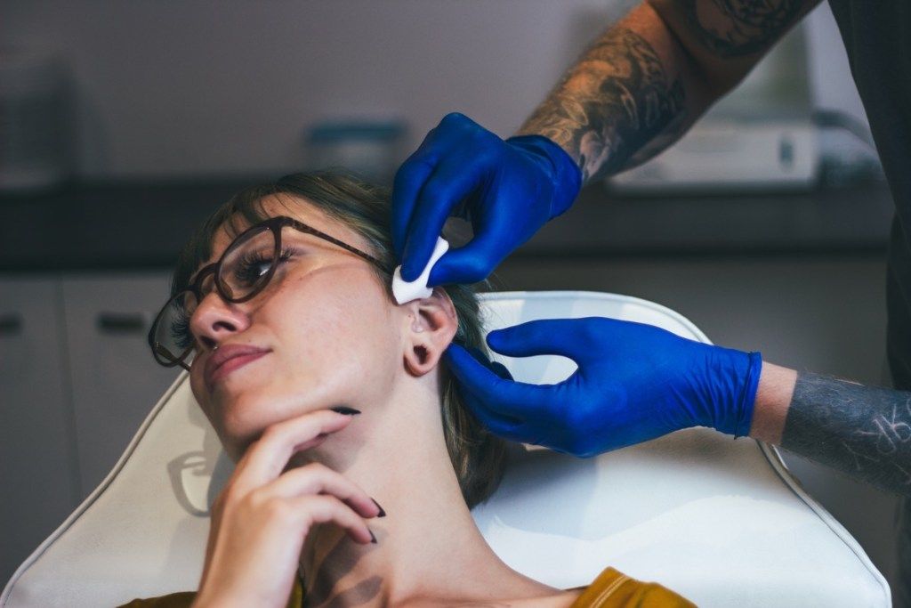 Mlada žena koja joj je probila uho. Čovjek prikazuje postupak piercinga sterilnom medicinskom opremom i rukavicama od lateksa. Postupak probijanja tijela (Mlada žena probušila uho. Čovjek pokazuje postupak piercinga sterilnom medicinskom opremom