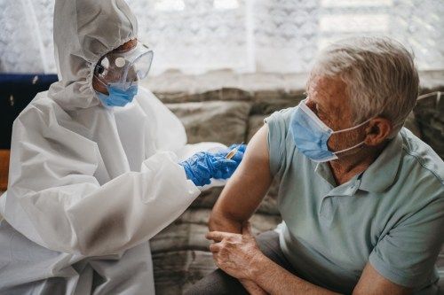 Zdravnica v zaščitni obleki daje cepivo proti virusu covid19 starejšemu moškemu doma