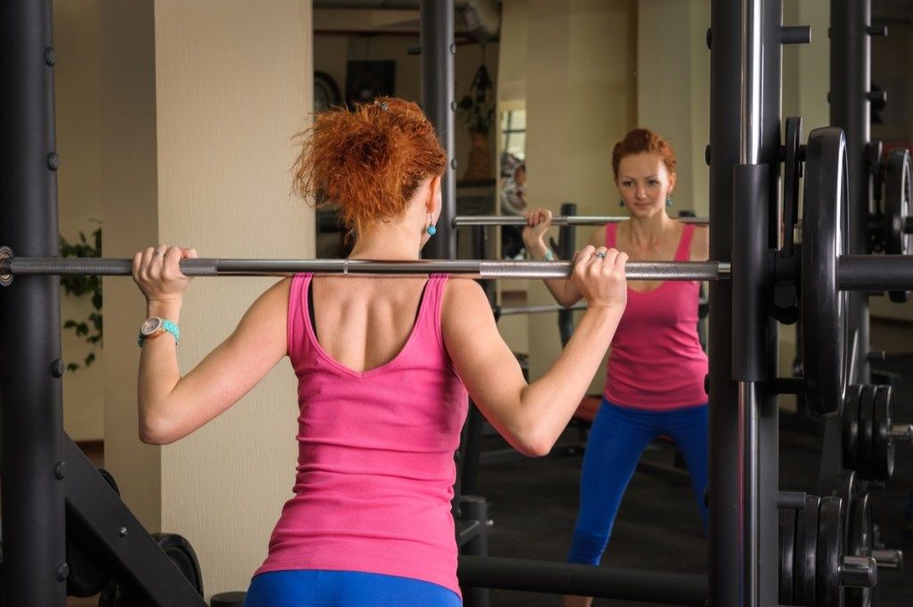 鏡の前でスミスマシンでバーベルとスクワットをしている若い赤い髪の少女