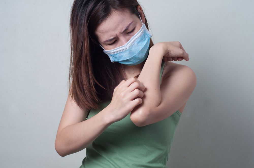 млада азиатска жена, която се почесва по ръката, докато носи хирургическа маска
