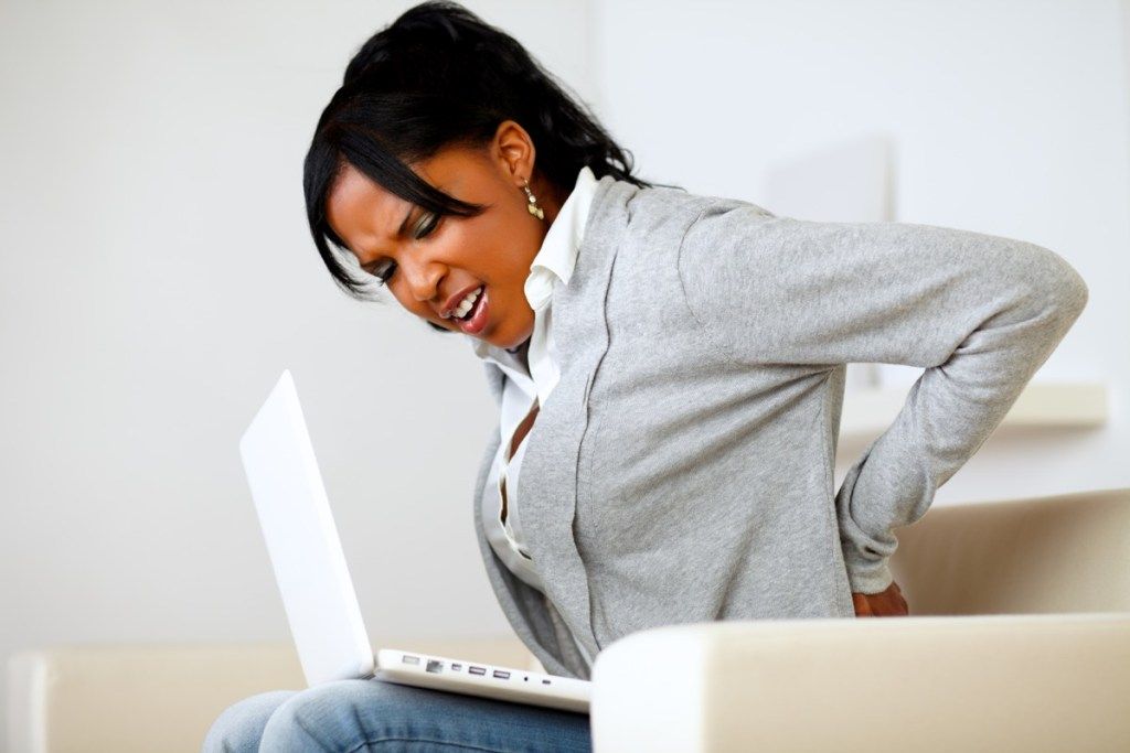 Frau, die Rückenschmerzen beim Sitzen auf einer Couch auf ihrem Laptop erlebt