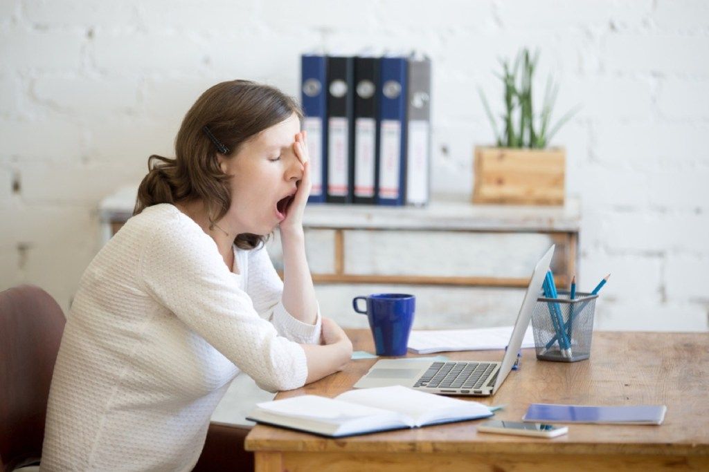 jovem mulher branca bocejando em sua mesa na frente de um laptop