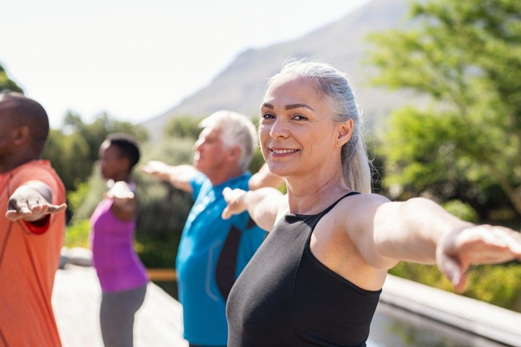Portret de femeie senior fericită care practică yoga în aer liber cu clasa de fitness. Femeie matură frumoasă care își întinde brațele și se uită la camera în aer liber. Portret de doamnă senină zâmbitoare cu brațele întinse în parc. (Portret de femeie senioră fericită pract