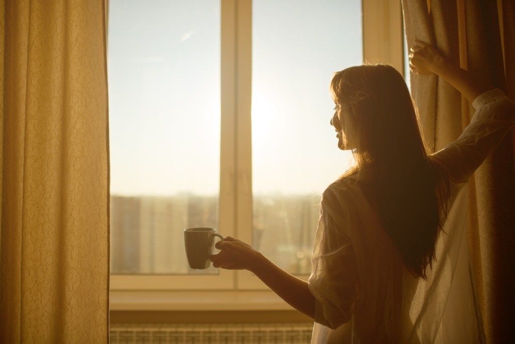 Kvinne som åpner gardiner om morgenen med kaffe