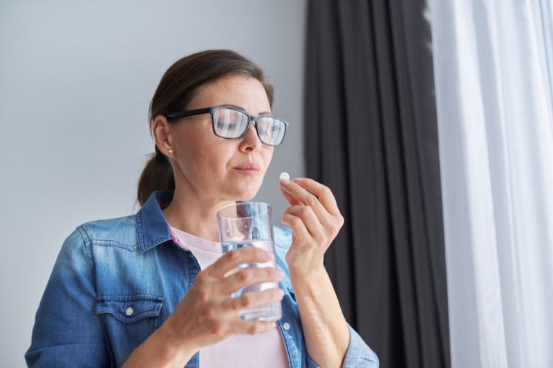   Femeie matură de vârstă mijlocie în haine casual acasă ținând pastilă și pahar cu apă proaspătă