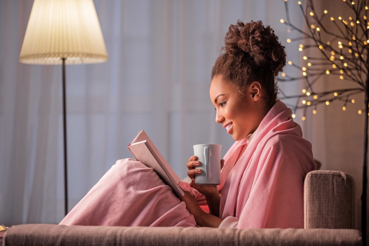 امرأة سوداء شابة تشرب الشاي وتقرأ كتابًا في رداء على الأريكة