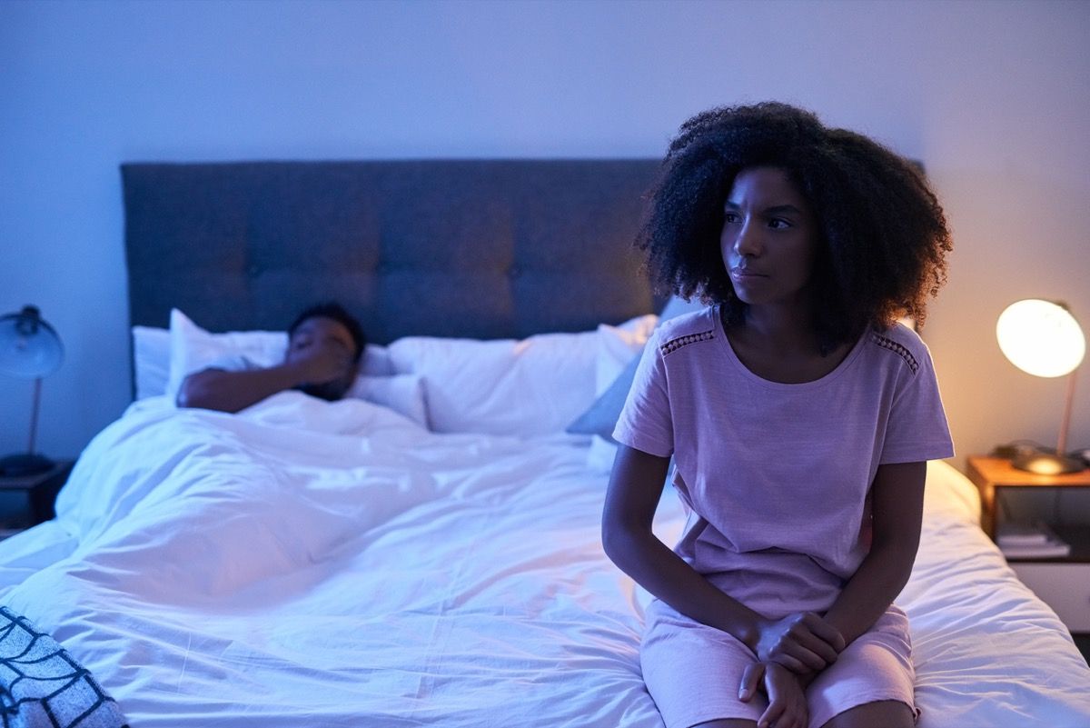 mladá černoška sedět strach v posteli s partnerem v pozadí
