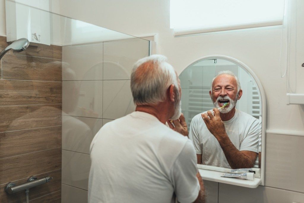 बूढ़ा आदमी आईने में दांत साफ करता है, दांतों को नुकसान पहुंचाता है