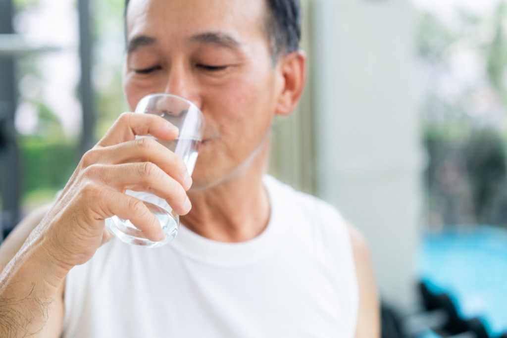 Hög man dricker mineralvatten i idrottshallkonditionmitten efter övning. Äldre hälsosam livsstil.