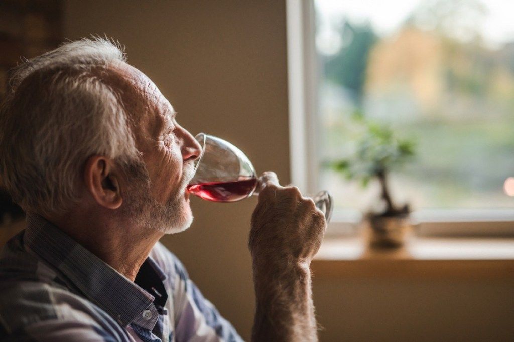 Zamyslený starší muž, ktorý doma pije červené víno a odvracia zrak