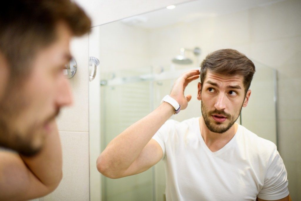 Jeune homme dans la salle de bain regardant dans le miroir et fixant ses cheveux avec des pellicules