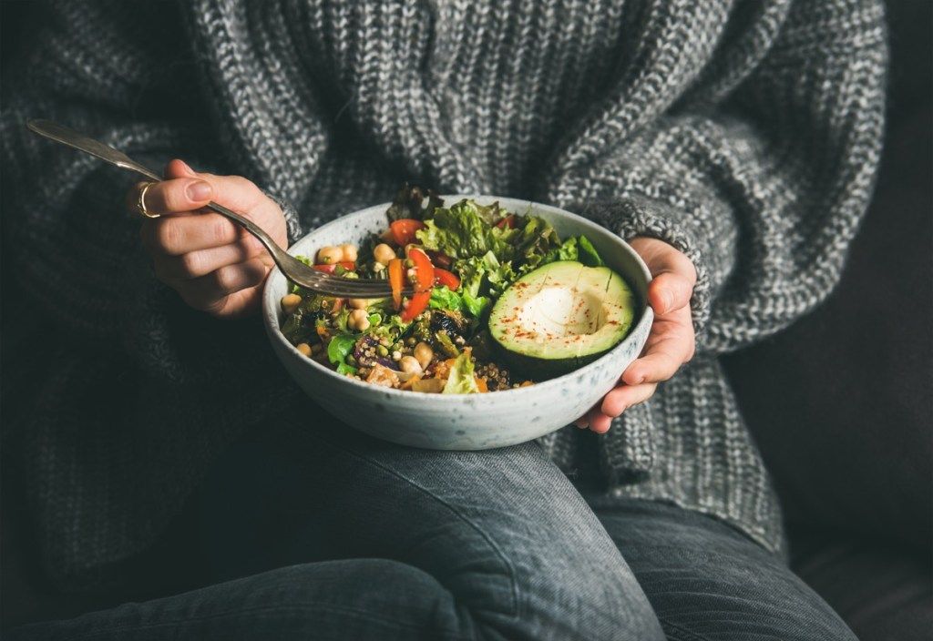 Cena vegetariana saludable. Mujer en jeans grises y suéter comiendo ensalada fresca, medio aguacate, granos, frijoles, verduras asadas del tazón de fuente de Buda