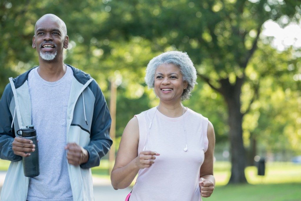 Vanhempi aikuinen afrikkalainen amerikkalainen pariskunta hymyilee ja lenkkeilee yhdessä julkisessa puistossa aurinkoisena päivänä. Aviomies ja vaimo käyttävät urheiluvaatteita.