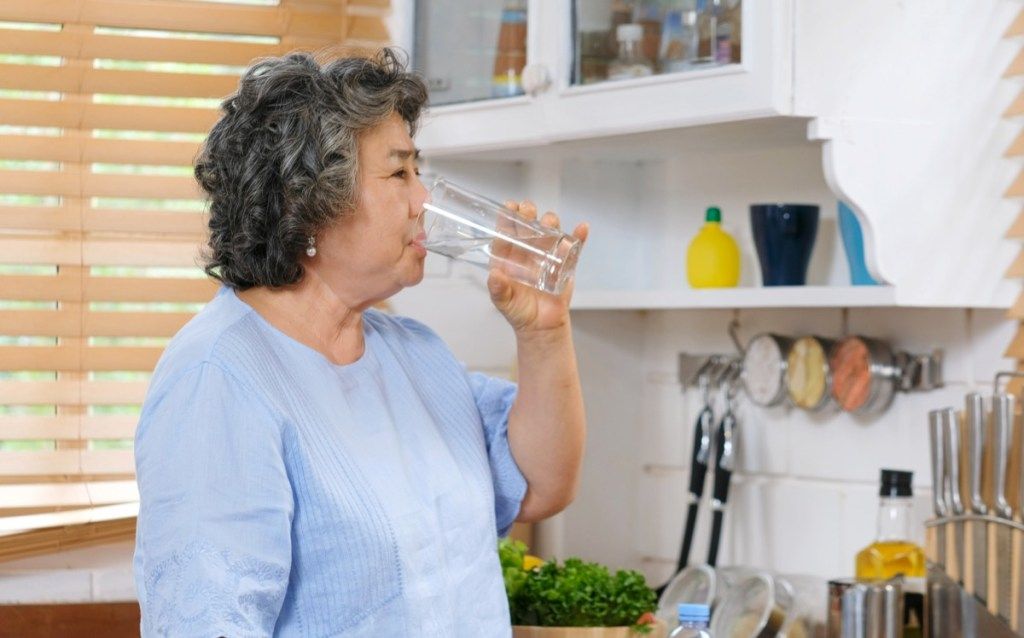 Idősebb ázsiai nő iszik vizet a konyhában