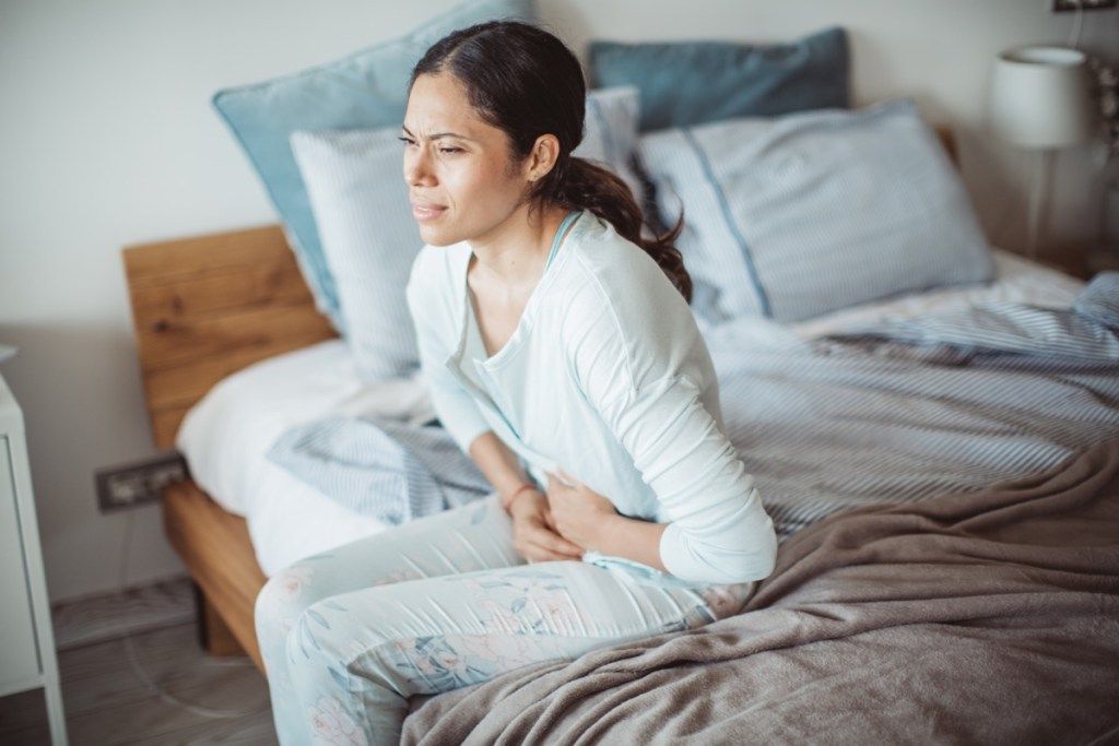 عورت بستر کے کنارے بیٹھی درد میں اپنا پیٹ تھام رہی ہے