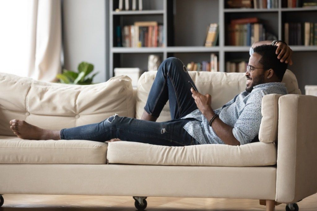 Täyspitkä rento nuori mies lasissa makaa mukavalla sohvalla nauttien viettää viikonlopun vapaa-aikaa chattailemalla sosiaalisessa mediassa puhelimessa ystävien kanssa, katsellen hauskoja videoita.