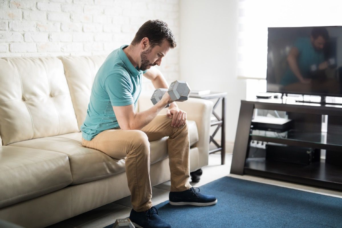hvid mand løfter vægte, mens han sidder på sin sofa foran tv