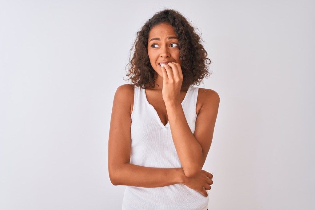 אישה ברזילאית צעירה לובשת חולצה מזדמנת עומדת על רקע לבן מבודד נראית לחוצה ועצבנית עם הידיים על הפה נושכות ציפורניים. בעיית חרדה.