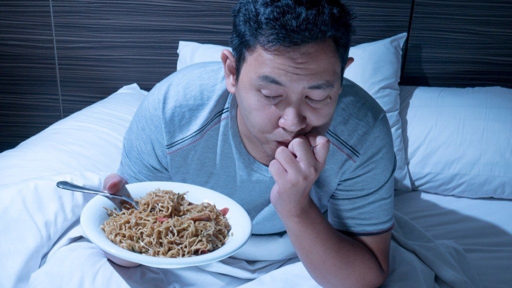 アジア人男性が夜遅くにベッドに座って麺のプレートを持って、40歳以降の健康に関する質問