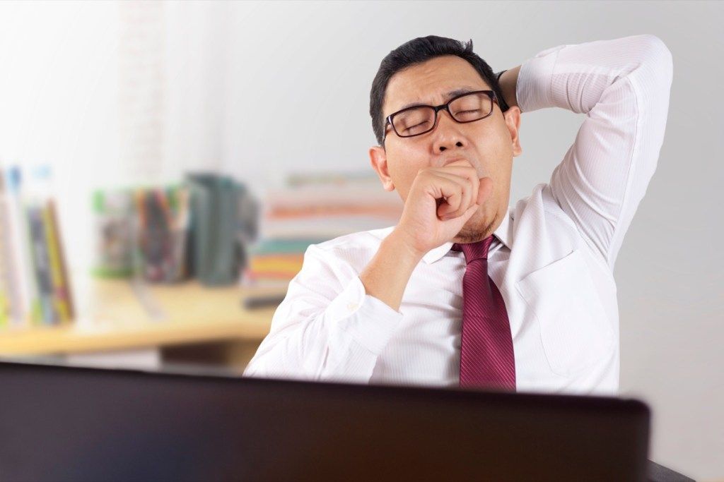 Hombre asiático sentado en su escritorio en el trabajo bostezando {síntomas de alergia}