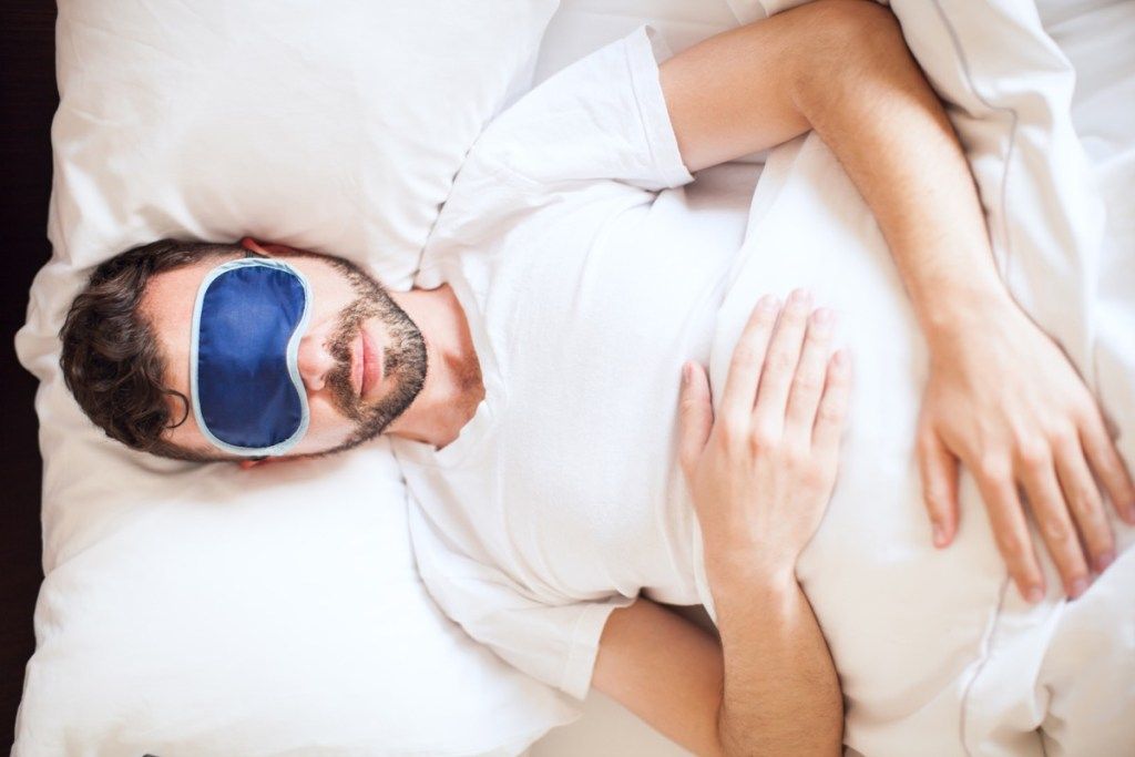 Vedere de sus a unui tânăr purtând o mască de somn în timp ce se odihnea în dormitor