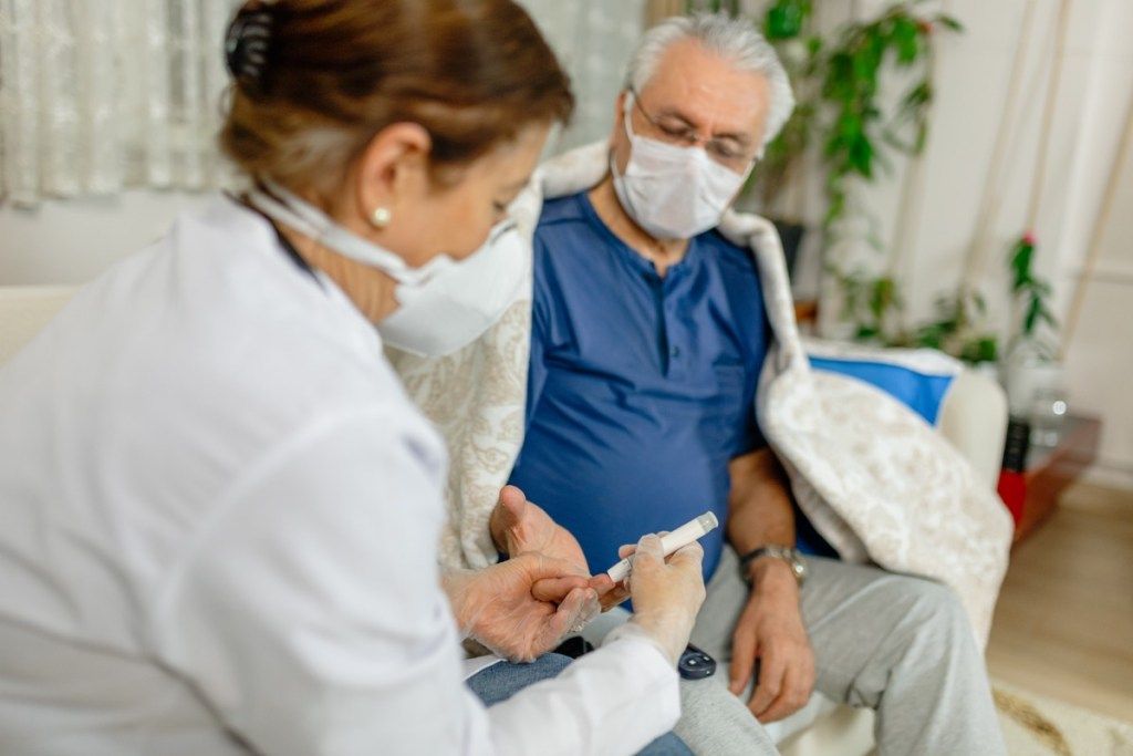 мъжът проверява нивата на кръвната захар от медицинска сестра, и двамата носят маски