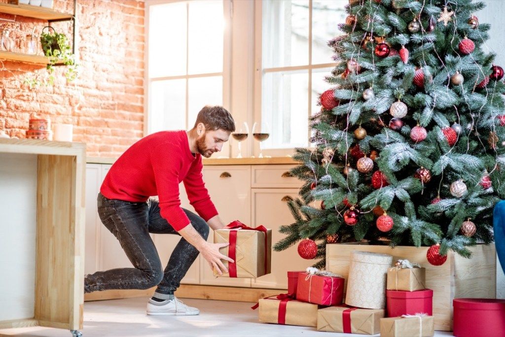 Hombre poniendo regalos bajo el árbol de Navidad preparándose para unas vacaciones de año nuevo en casa