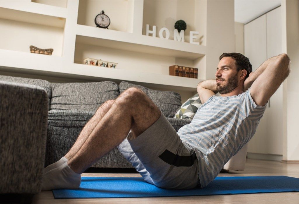 एथलेटिक आदमी घर पर व्यायाम की चटाई पर बैठा है और अपने सिर के पीछे हाथों से बैठकर व्यायाम करता है।