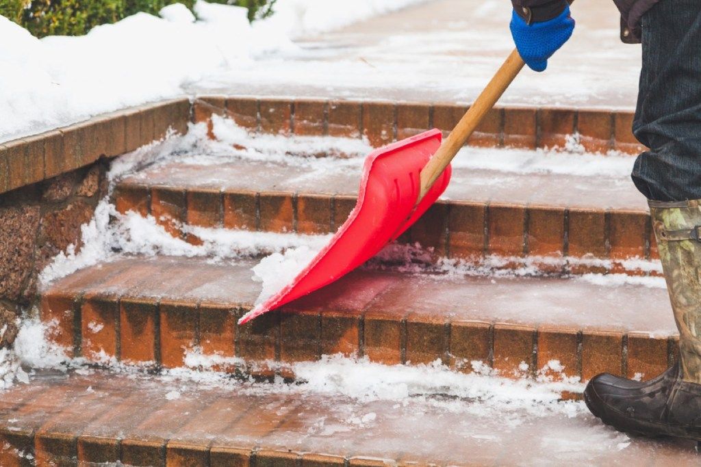 pessoa removendo neve em degraus de tijolos com uma pá vermelha