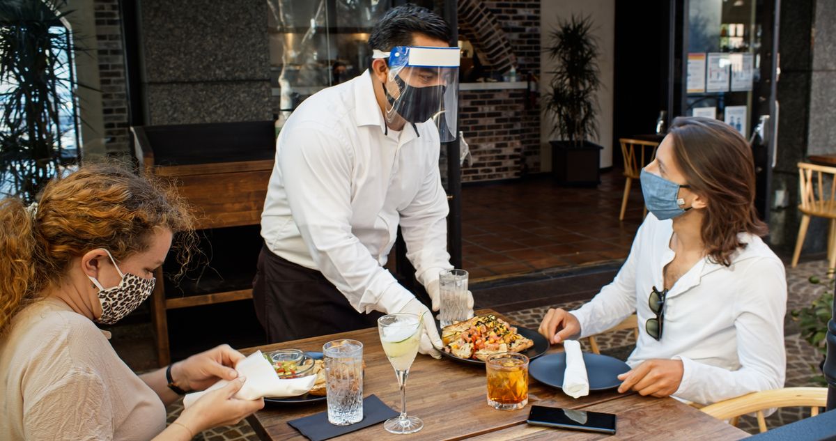 Un camarero de un restaurante de lujo que lleva una mascarilla y un protector facial se acerca a la mesa con una bandeja, que sirve a las mujeres que están sentadas y llevan mascarillas.