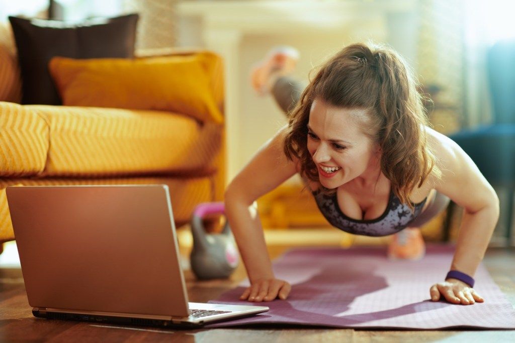 sorridente donna sana in abiti fitness nel soggiorno moderno guardando tutorial di fitness su internet tramite laptop e facendo flessioni sul tappetino fitness. (sorridente donna in buona salute in abiti da fitness nel soggiorno moderno guardando il tutorial di fitness su inte