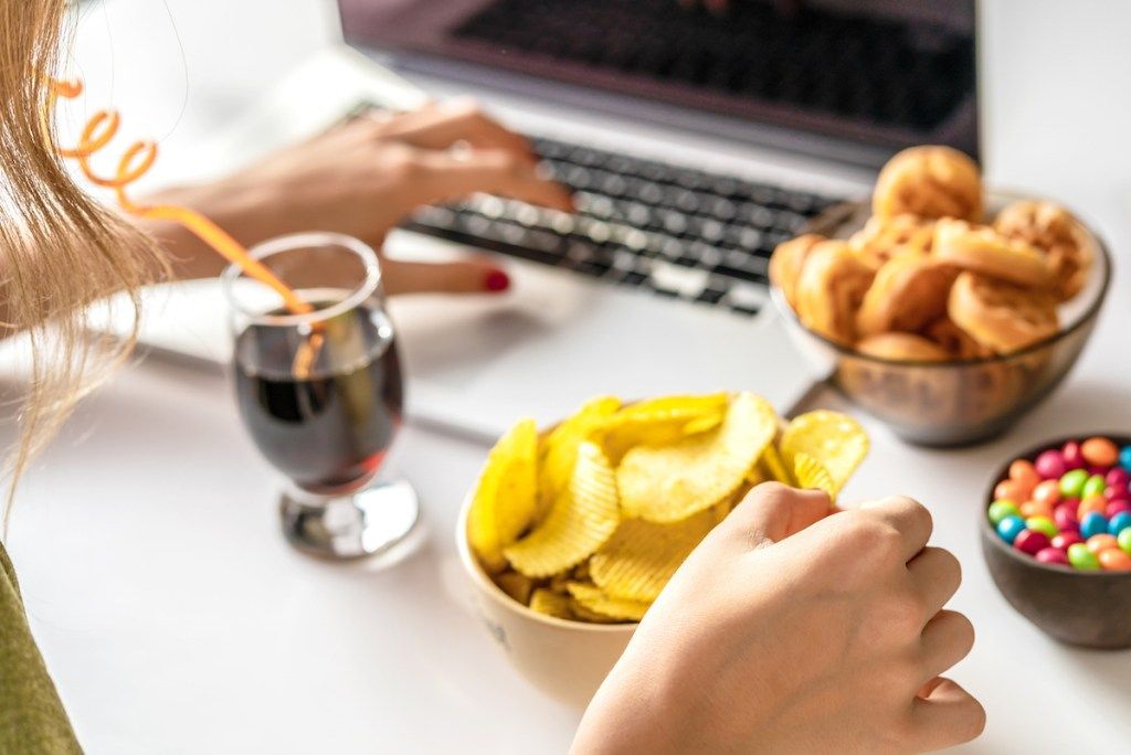 여자는 컴퓨터에서 일하고 건강에 해로운 음식을 먹는다 : 칩, 크래커, 사탕, 와플, 소다