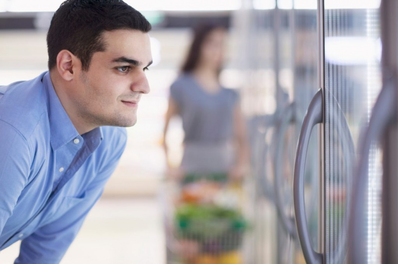   Seorang lelaki melihat melalui pintu penyejuk atau peti sejuk semasa membeli-belah di bahagian makanan sejuk atau sejuk beku di kedai