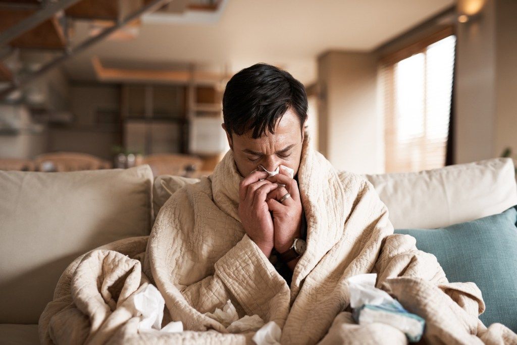 Antklodėje įsisupęs vyras sėdi ant sofos ir pučia nosį su gripo simptomais, nes gripo sezonas sutampa su koronaviruso pandemija.