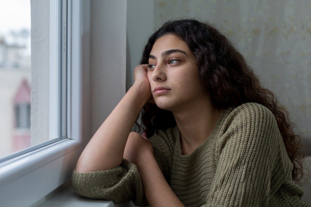 Teini-ikäinen tyttö, jolla on tummat hiukset, näyttää ikkunasta surullisella ilmeellä.