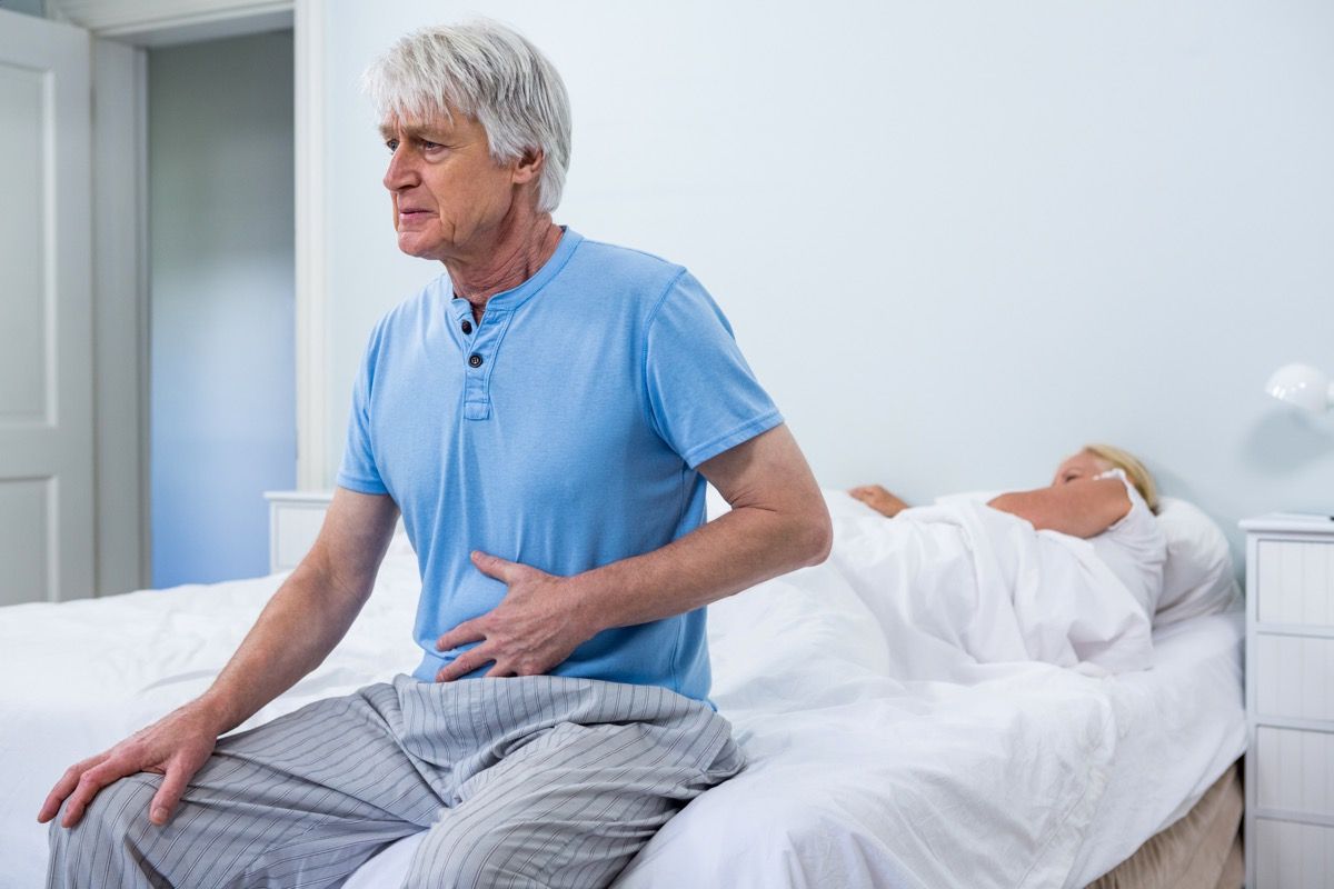 vyresnysis vyras su baltais plaukais sėdi lovos krašte, laikydamas pilvą, o žmona miega, ką gali reikšti jūsų pilvo skausmas