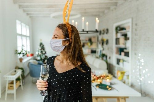 Hình ảnh một phụ nữ trẻ cầm ly sâm panh, đeo mặt nạ bảo hộ và đeo gạc tuần lộc ăn mừng và tổ chức bữa tiệc tối Giáng sinh tại nhà trong đại dịch COVID-19.