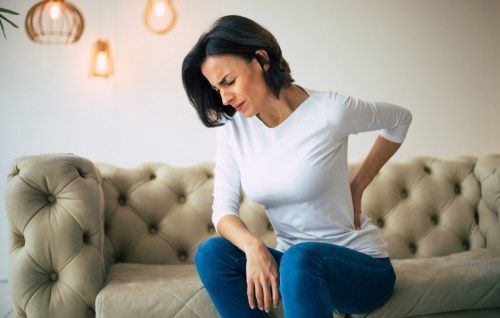 Aksialna bolečina. Fotografija od blizu prizadete ženske, ki sedi na kavču in jo z levo drži spodnji del hrbta.