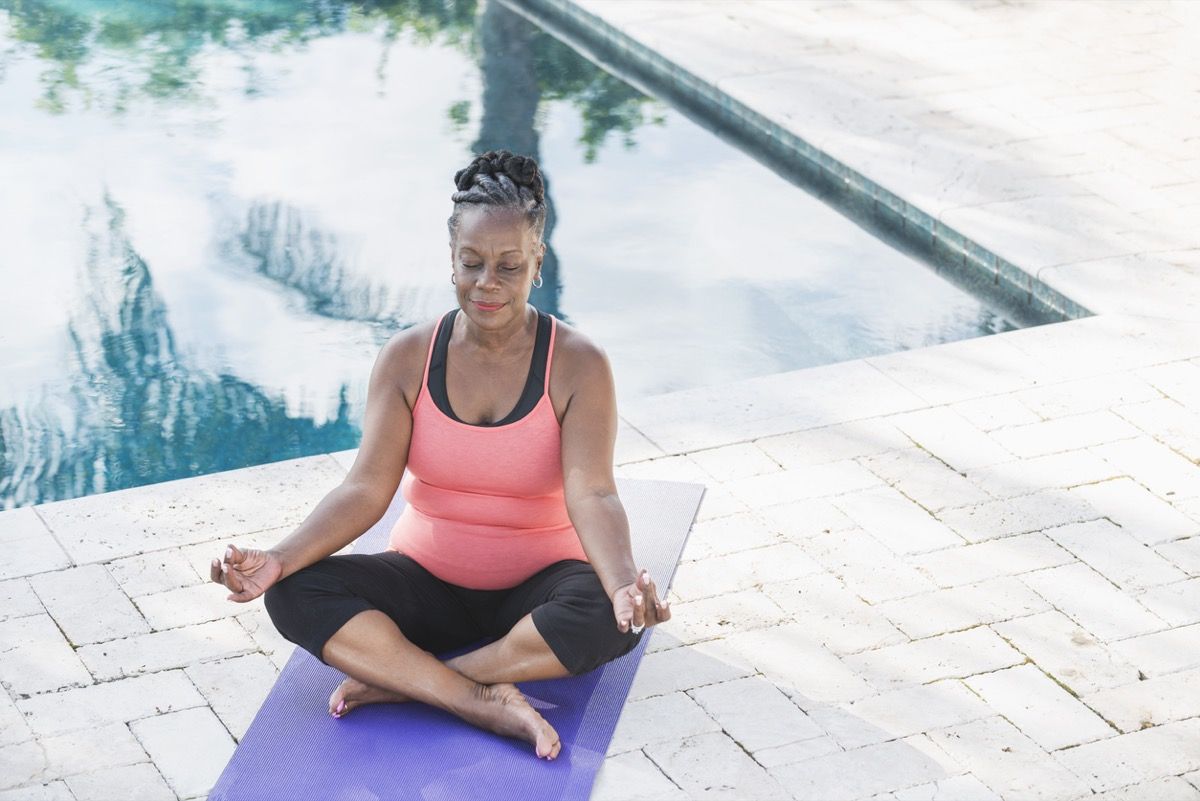 по-възрастна чернокожа жена, медитираща върху йога постелка край басейн