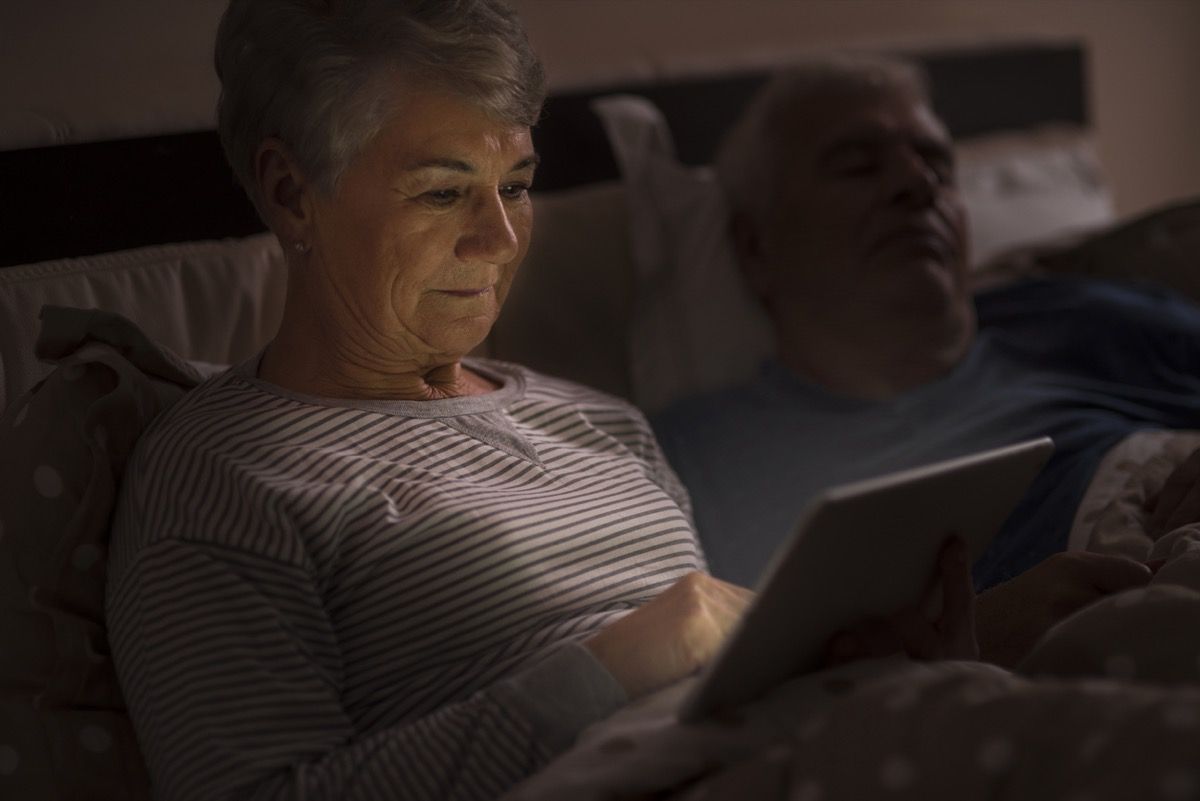 по-възрастна бяла жена, използваща ipad в леглото, докато съпругът спи до нея