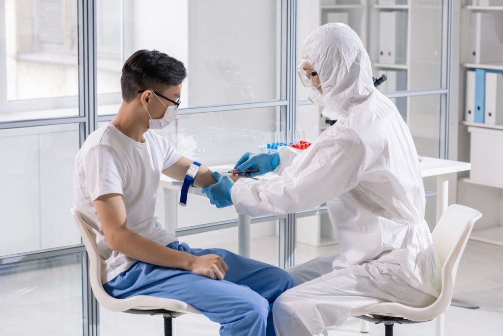 Jaunas vyras kaukėje, sėdintis laboratorijoje, o gydytojas su apsauginiais kombinezonais paimdamas kraują švirkšte analizei