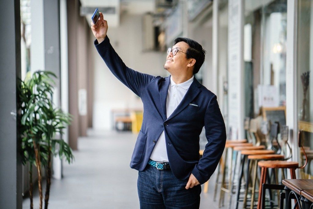 mies yllään sininen bleiseri ja tummat farkut, joilla on älypuhelin ja puhuu selfie