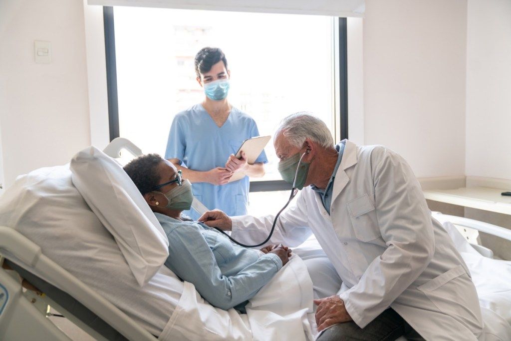 medic de sex masculin care verifică pacientul internat în spital care îi ascultă inima și asistenta care stă lângă ei, purtând măști de protecție - stiluri de viață pandemice