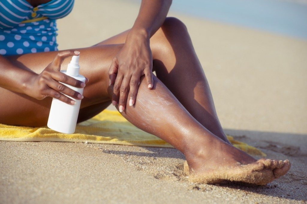 Mujer negra aplicando, rociando crema de protección solar en las piernas, hábitos después de los 40