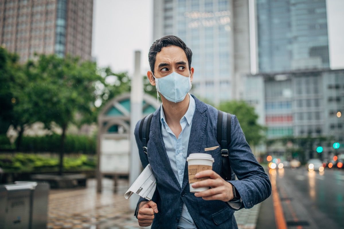 vīrietis uzvalkā, turēdams laikrakstu un kafiju un valkājot sejas masku pilsētā