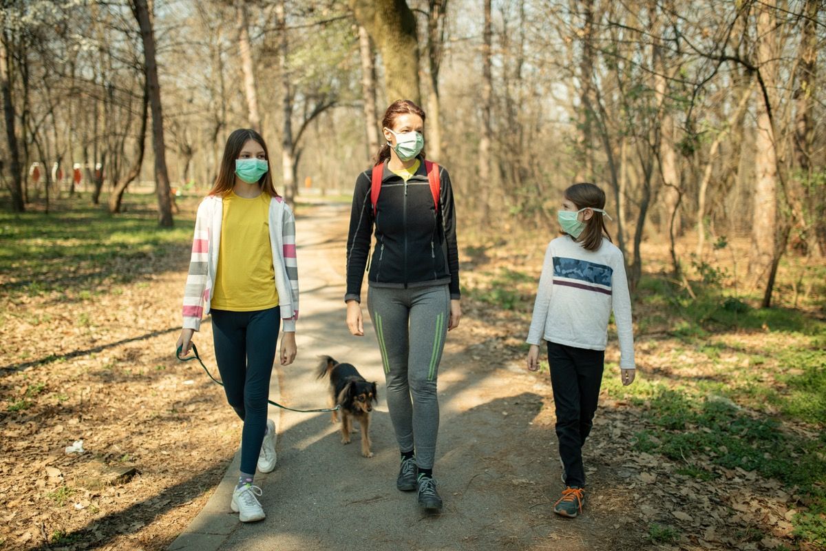 äiti ja kaksi tytärtä kasvonaamareissa kävelevät koiraa puistossa