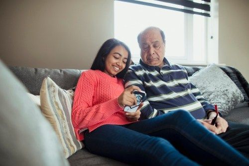 Una vista frontal de un abuelo sentado en un sofá con su nieta, la nieta joven está usando su teléfono para jugar un juego en línea.