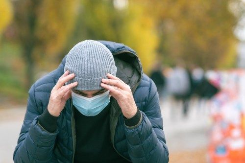 Un uomo con una maschera medica nel parco con la mano sulla testa a causa di un mal di testa.