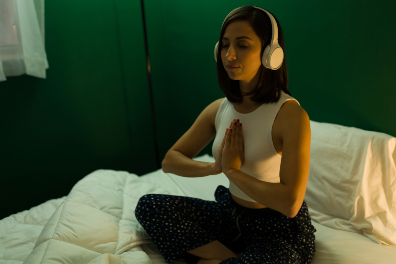   Nainen meditoi ja kuuntelee kuulokkeita sängyssä.